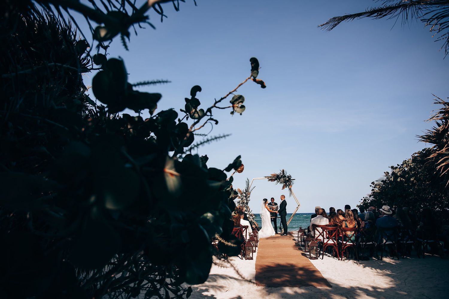 Beach wedding setting with silhouetted palm trees against bright blue sky.
