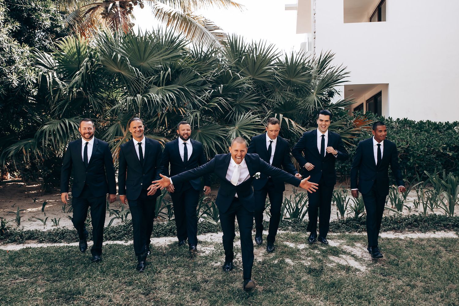 Groomsmen in black suits and ties stand in line against tropical palm garden backdrop.