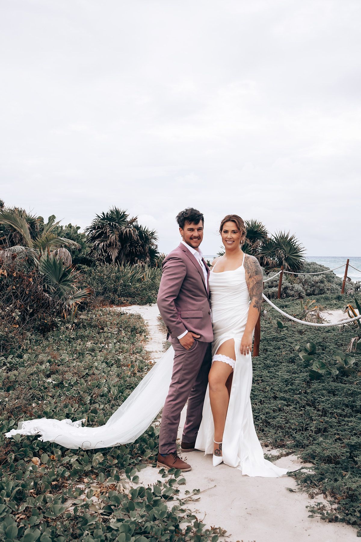 Wedding couple posing on a tropical beach path with flowing white dress and palm trees in the background.