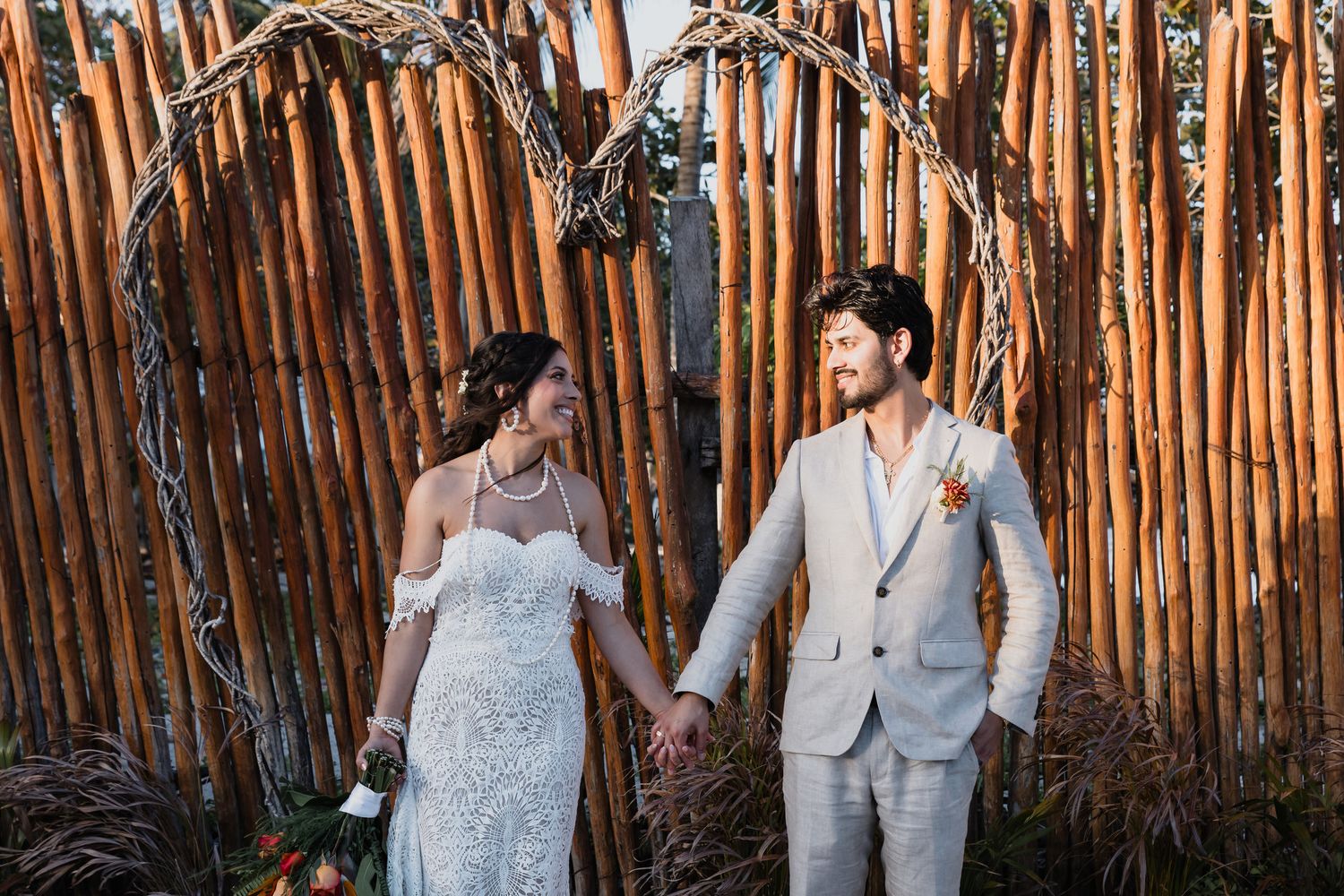 A bride and groom hold hands and smile at each other beneath a rustic wooden arch at golden hour.