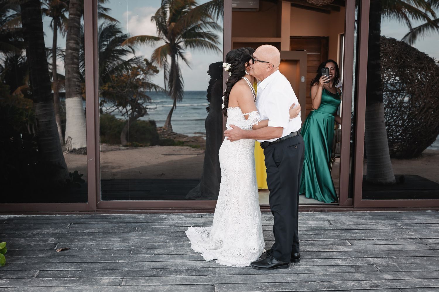 Emotional father-daughter first look embrace at beachside venue with palm trees and ocean in background.