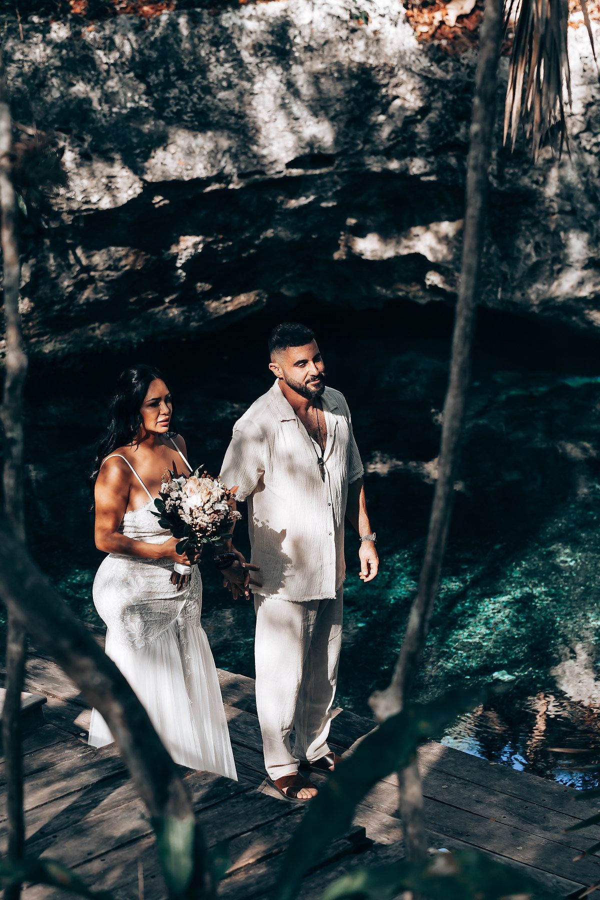 A couple in white attire poses in a dramatic forest setting with rocky cave formations in the background.