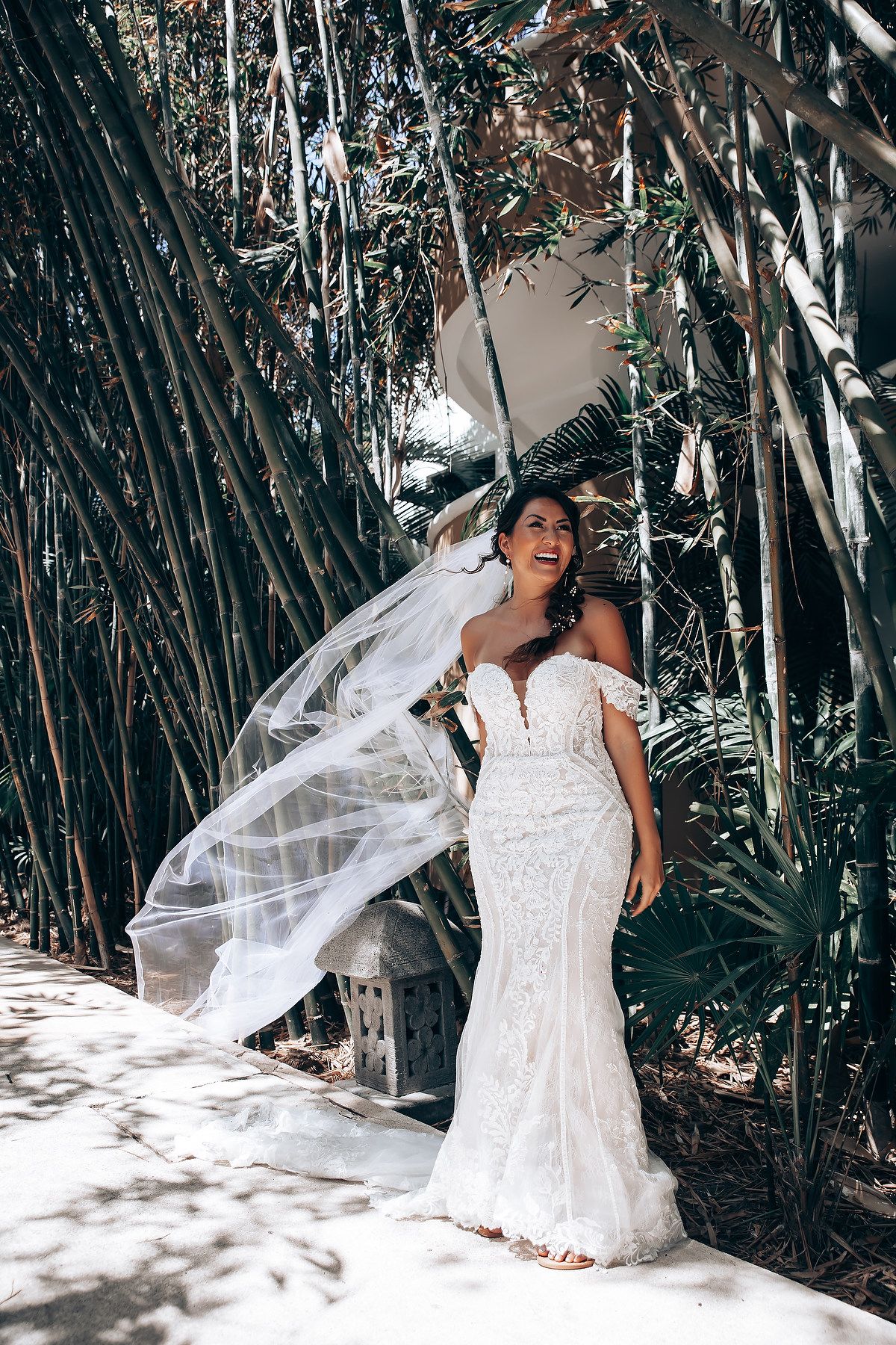 Bride in white lace dress poses playfully with flowing veil in tropical bamboo forest setting.