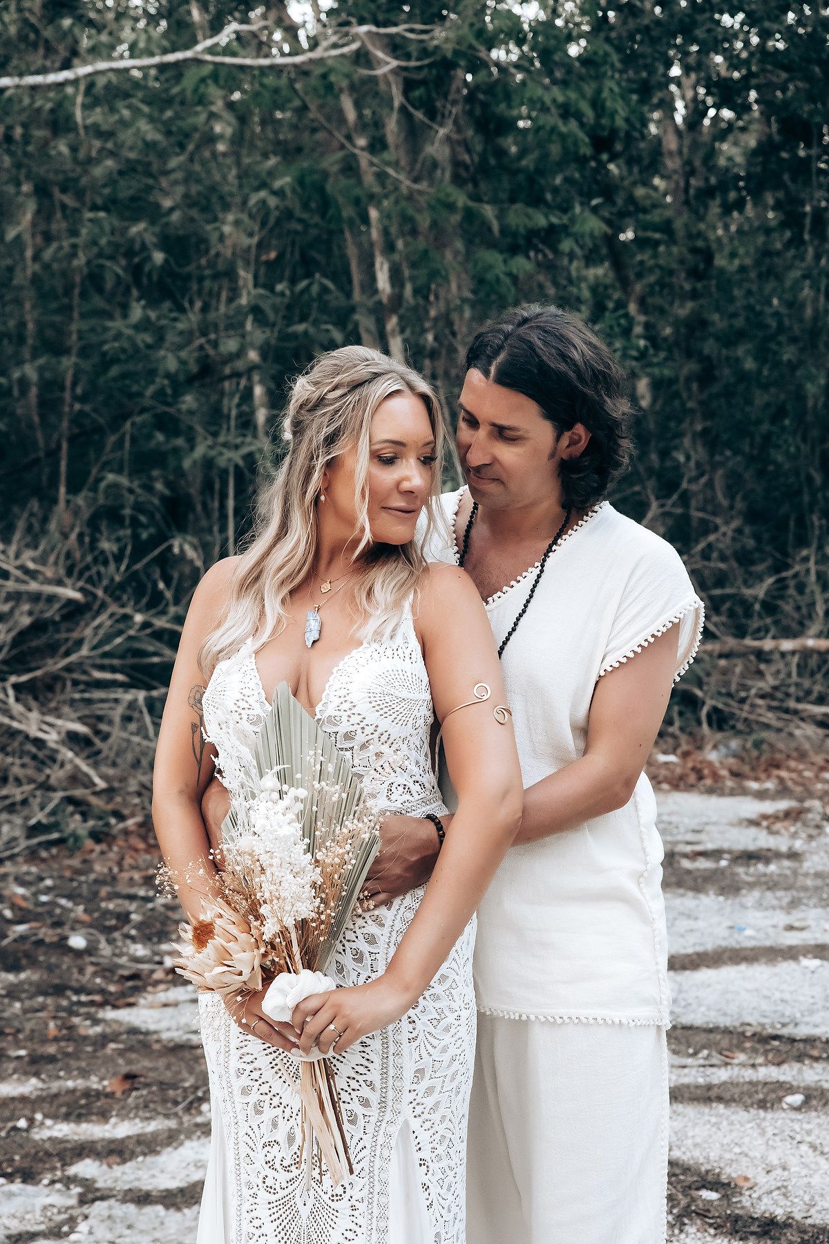 A romantic couple in flowing white outfits share an intimate moment among trees during a forest photoshoot.