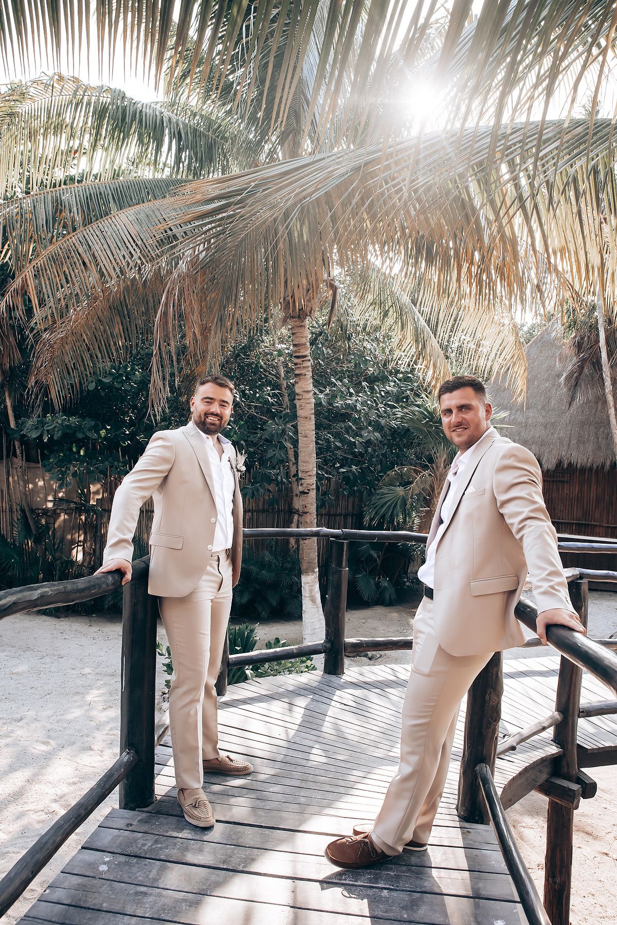 Two people in cream-colored suits stand on a wooden deck surrounded by tropical palm trees and sunlight.