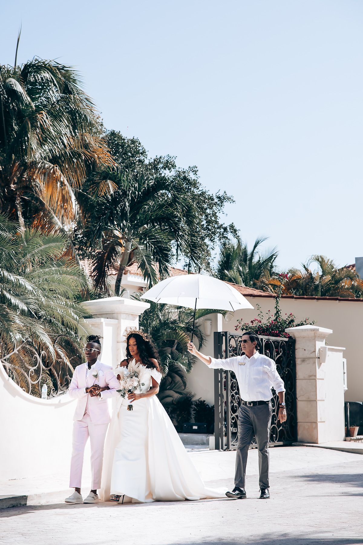 Wedding party in white and pink attire gathered on a sunny terrace with palm trees.