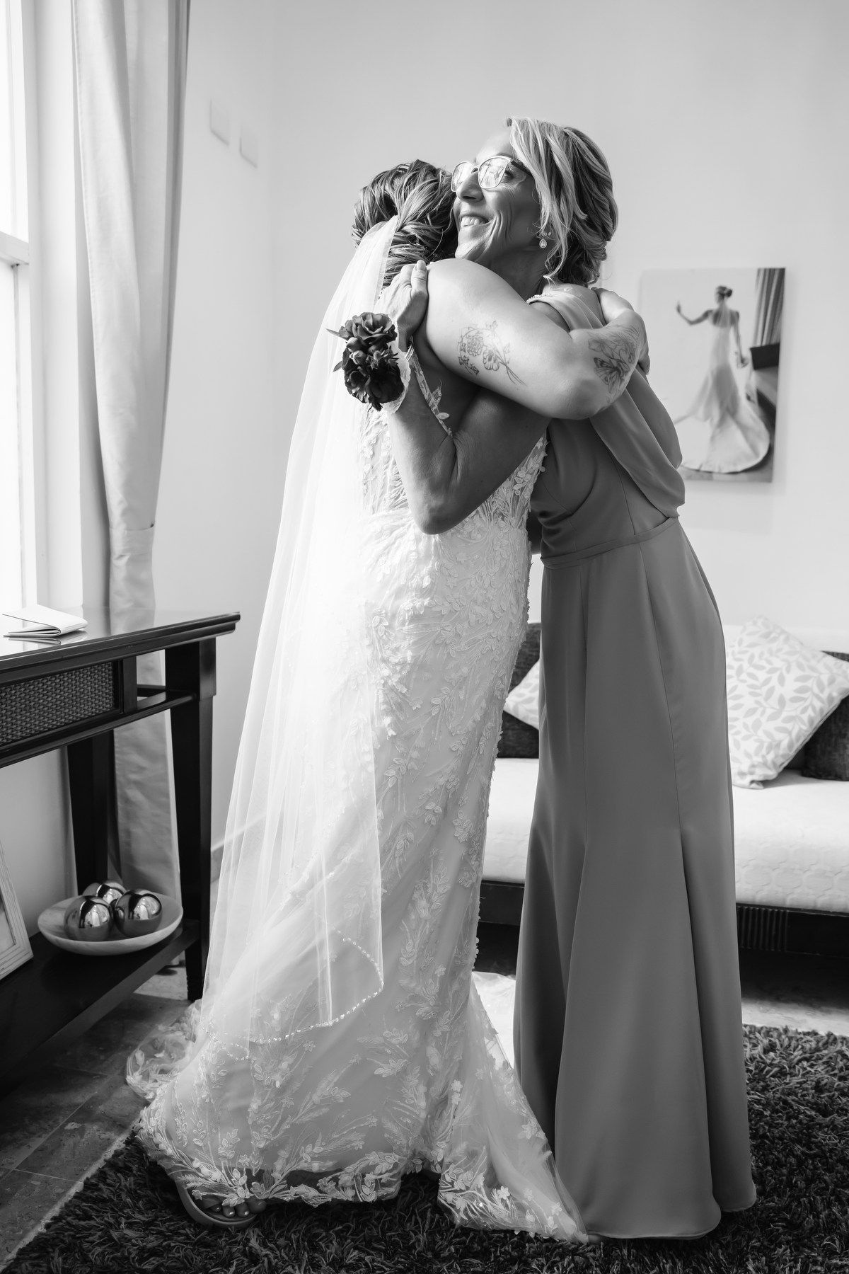 Black and white photo of bride in lace gown sharing an emotional hug with bridesmaid before wedding.