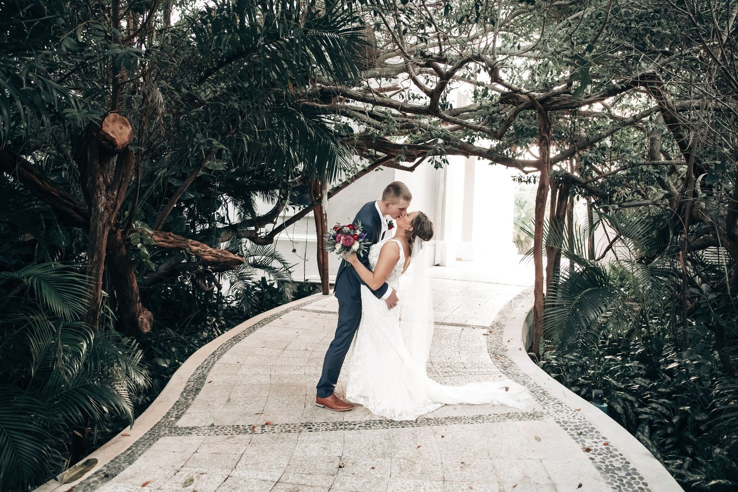 A bride and groom share a romantic kiss on a mosaic garden pathway surrounded by lush tropical greenery.