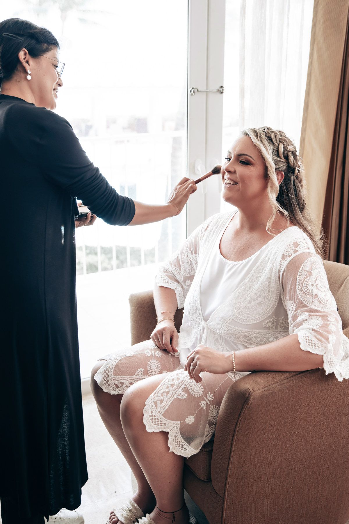 Makeup artist in black dress applying makeup to smiling bride in white lace dress seated in armchair at Dreams Tulum
