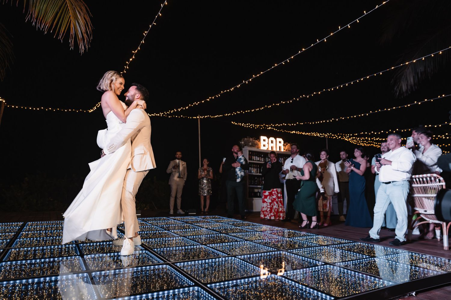 Groom lifting bride during first dance on sparkling LED dance floor at outdoor night wedding.