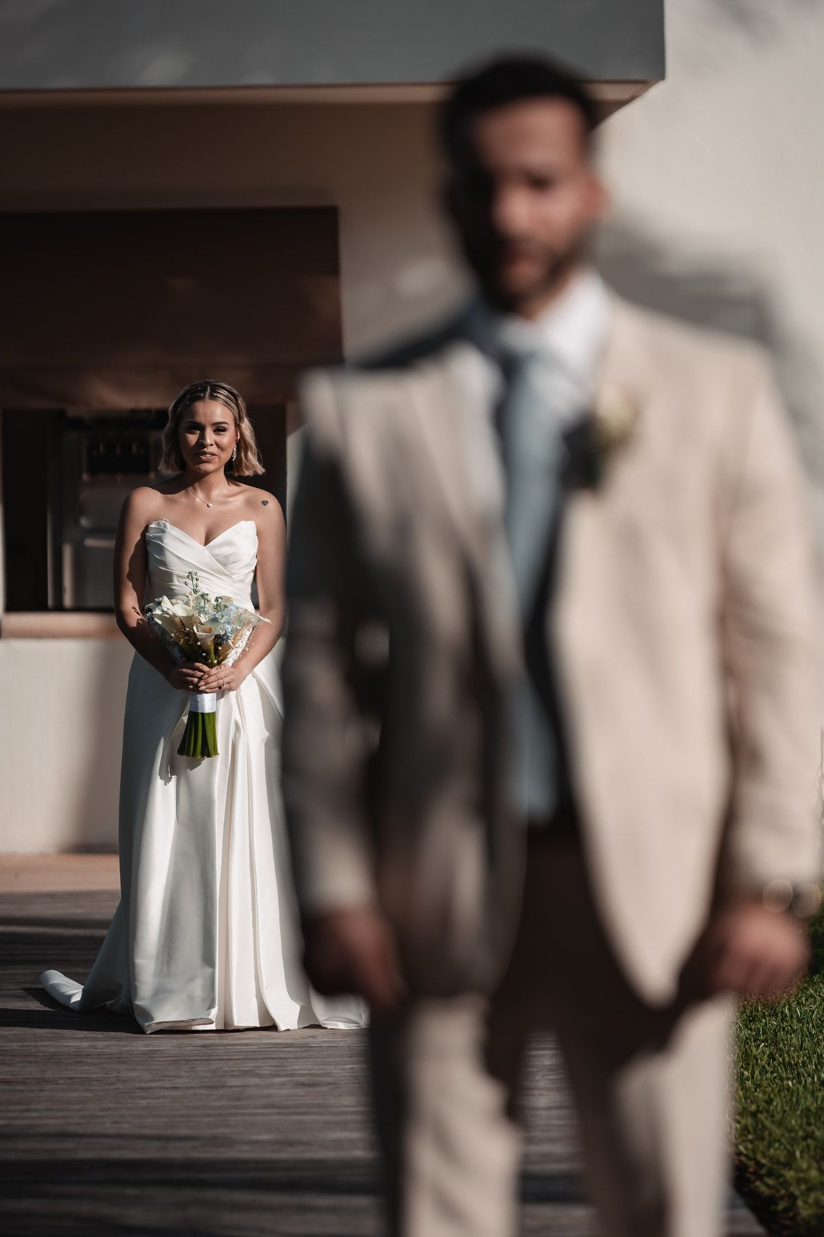 Bride in white gown holding bouquet approaching groom in beige suit.