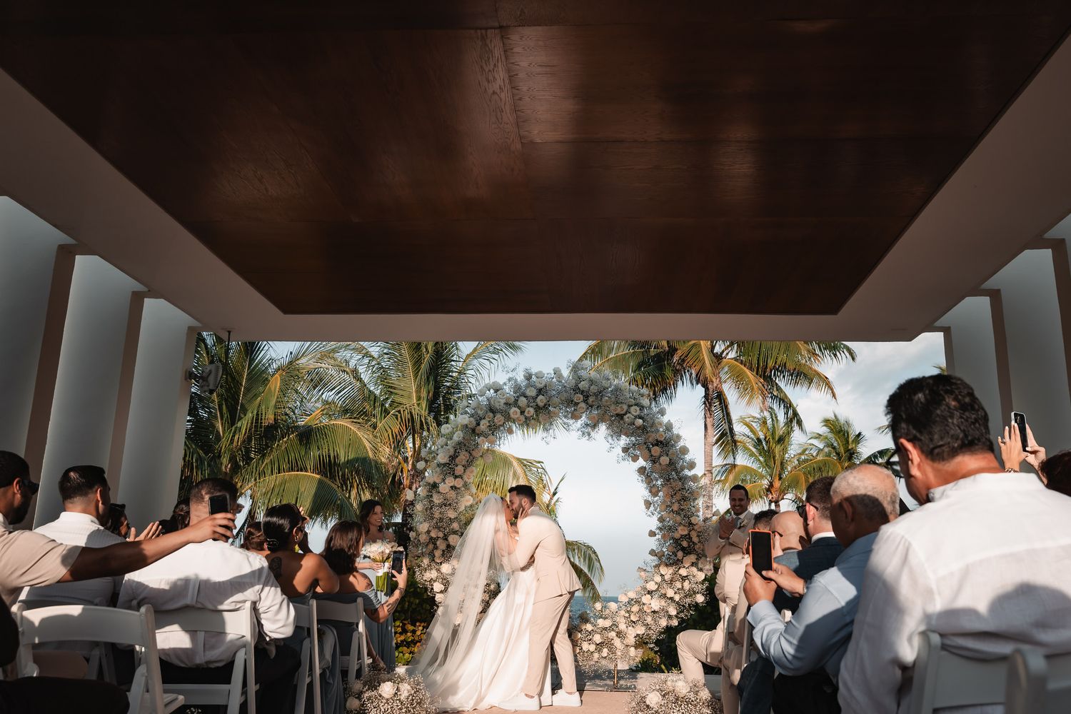 Bride and groom sharing a first kiss under a floral arch at an outdoor tropical wedding ceremony.