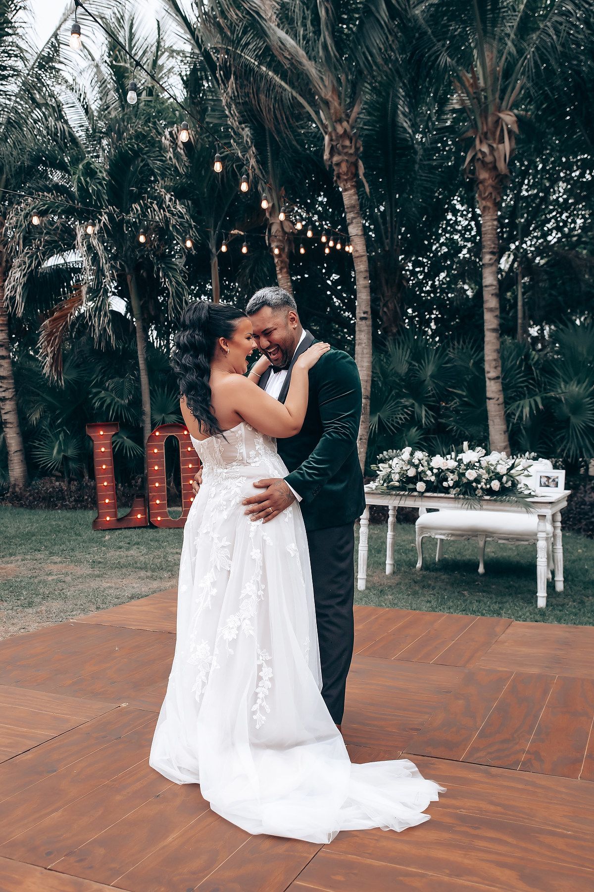 Newlyweds dance together on outdoor terrace surrounded by palm trees and string lights.