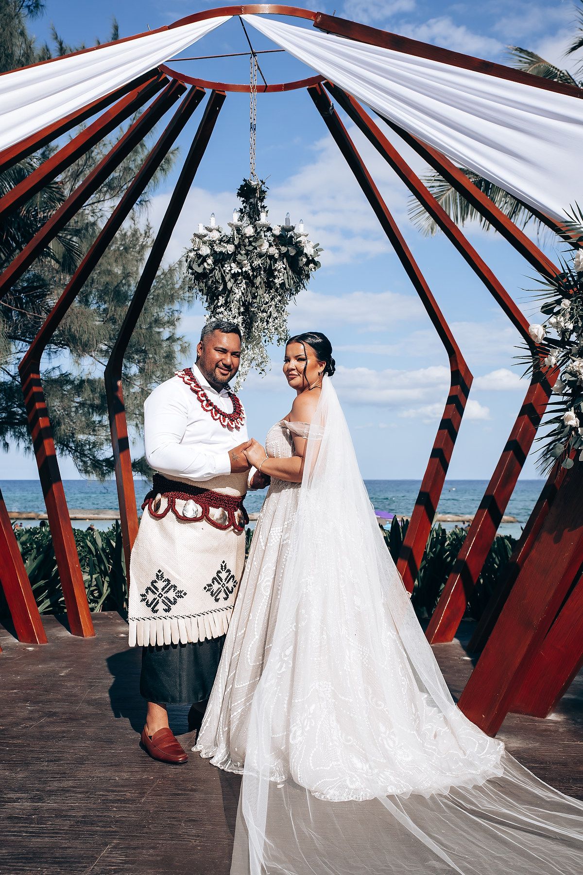 A couple poses in a red and white beachside gazebo structure during a tropical wedding photoshoot.