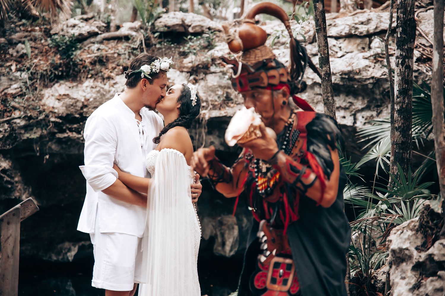 A couple shares an intimate moment in a wooded outdoor setting while a photographer captures the scene.