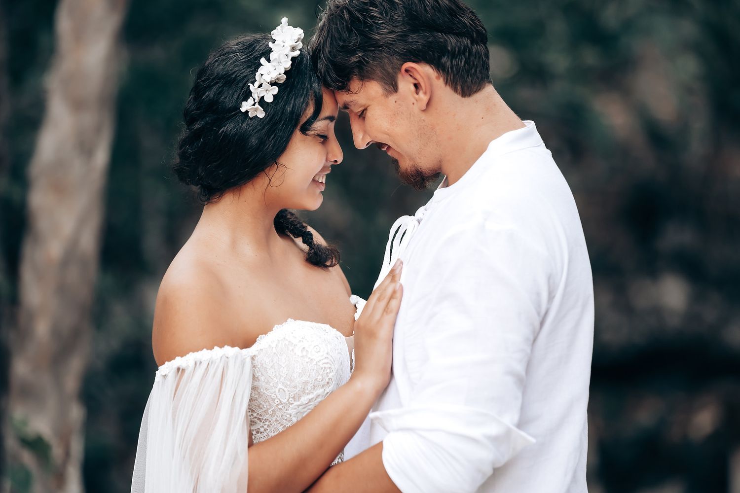 A tender moment captured between a couple wearing white as they embrace in a wooded outdoor setting.