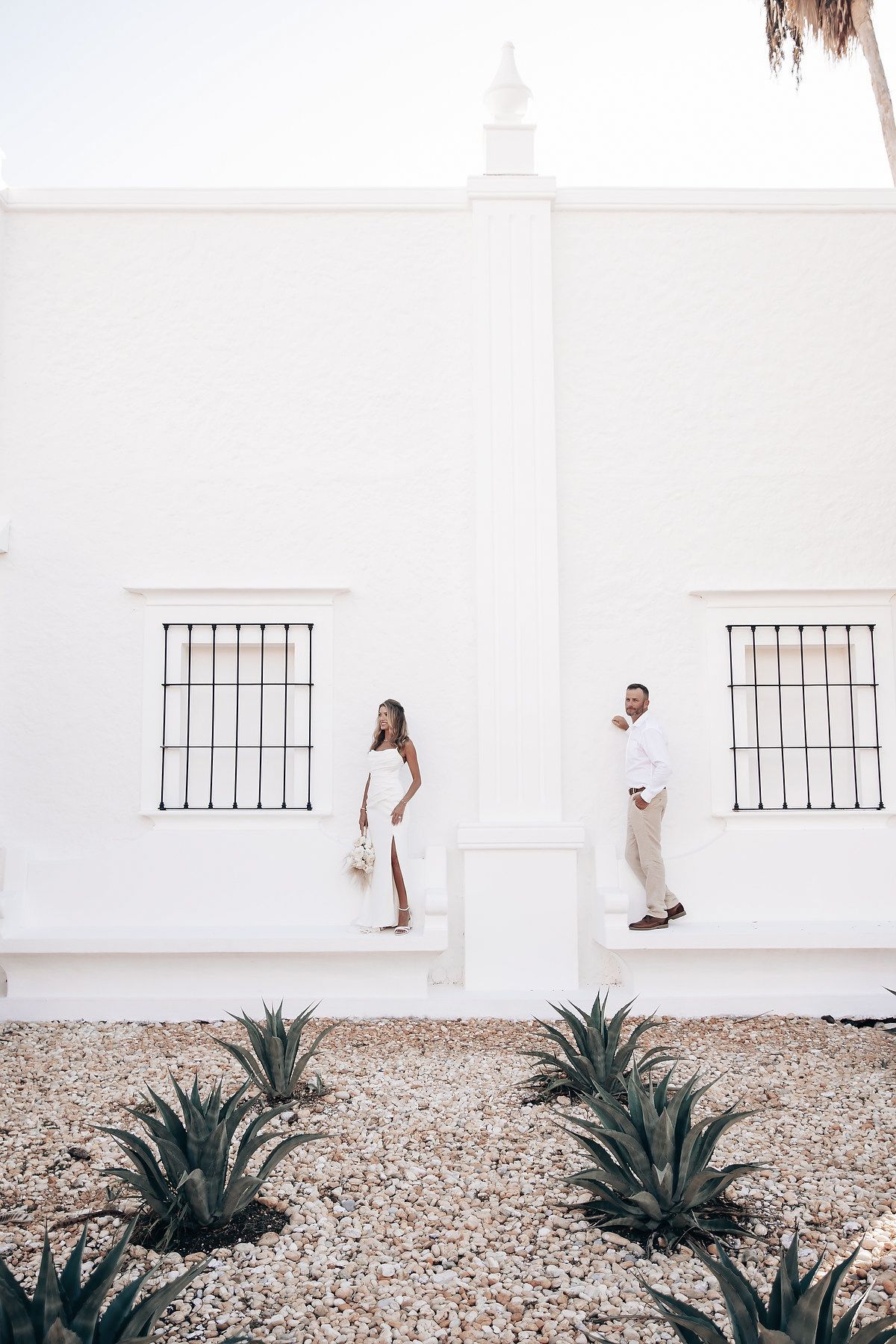 A couple stands in front of a white wall, with the woman wearing a long, flowing dress.