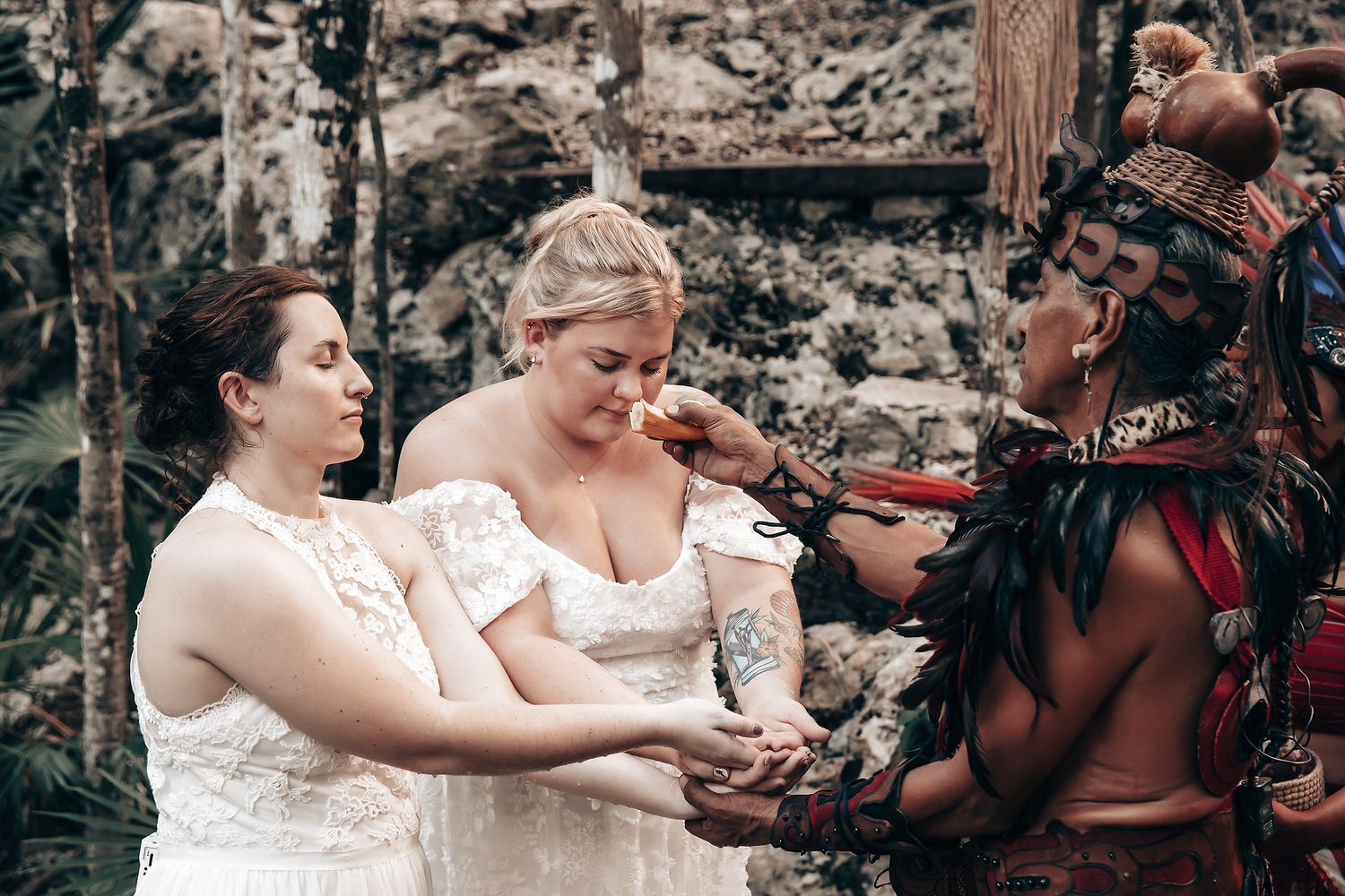 Two brides in white wedding dresses hold hands with a tribal man in a lush, outdoor setting.