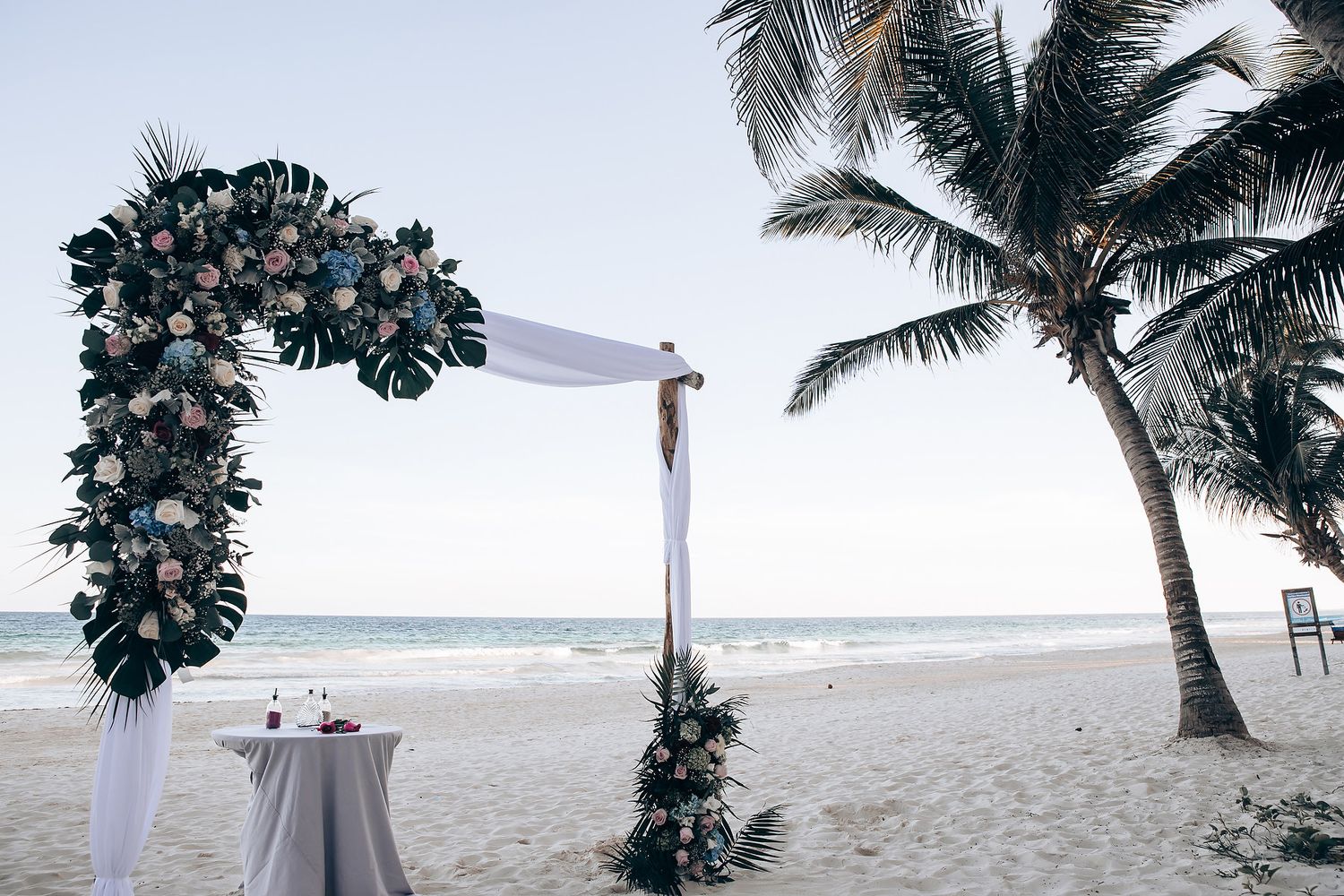A tropical beach wedding setup with a floral arch, palm trees, and a table on the sandy shore.
