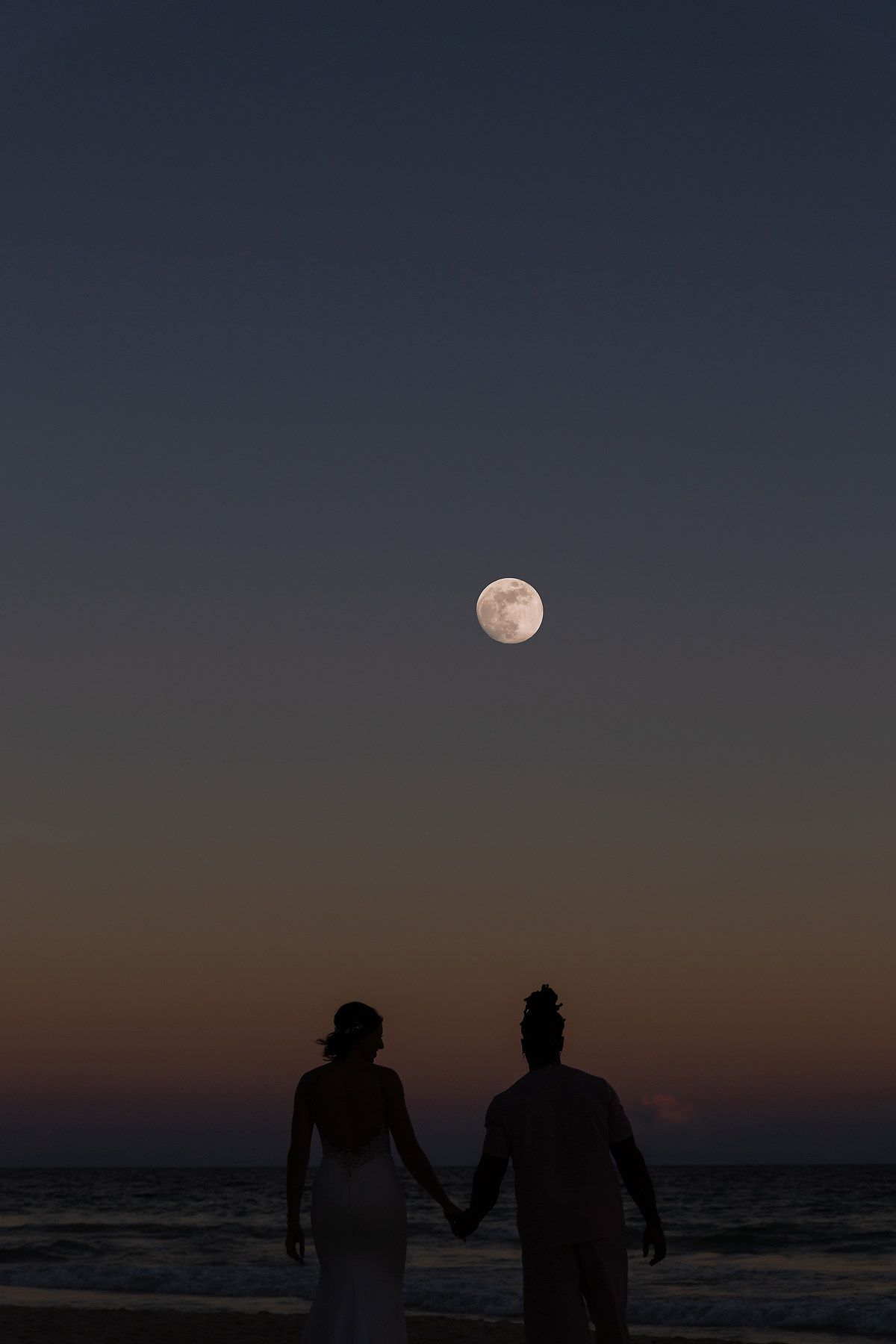 Two silhouetted figures holding hands while standing on a beach with a full moon in the sky.