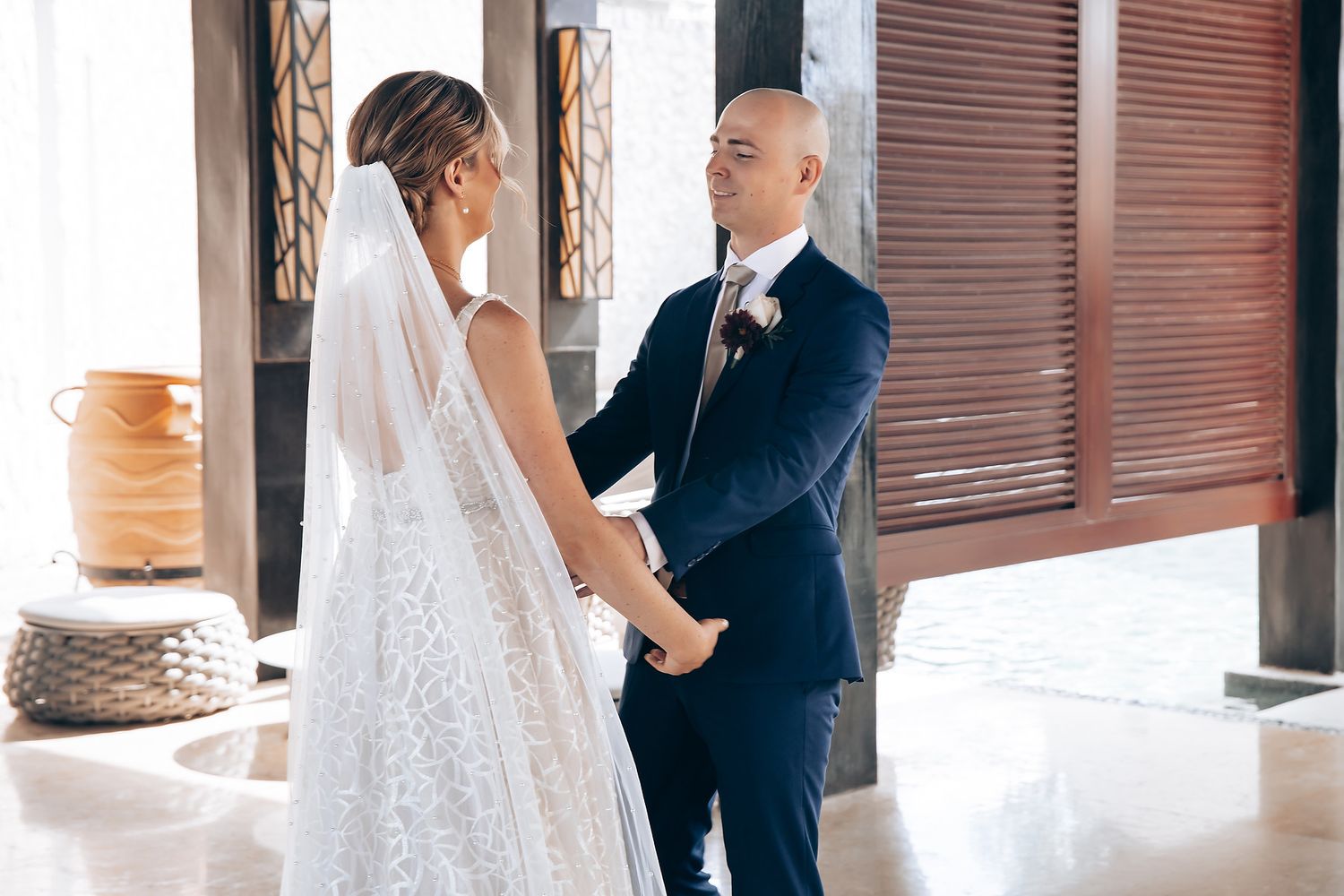 A bride and groom stand together in a doorway, the bride wearing a long, lace wedding dress.