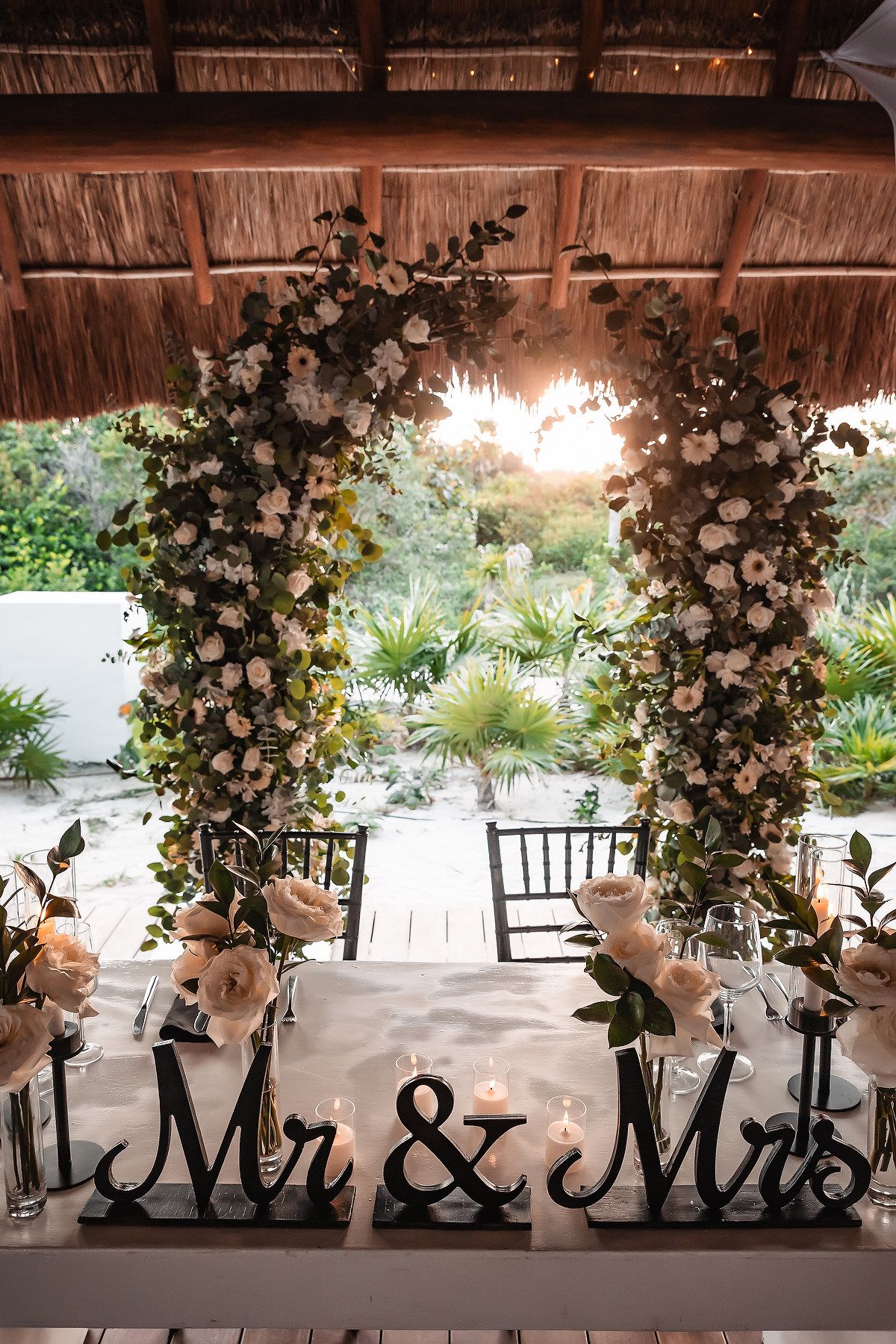 Elegant wedding sweetheart table with Mr & Mrs signs, white floral arch, candles, under a thatched roof with tropical garden views.