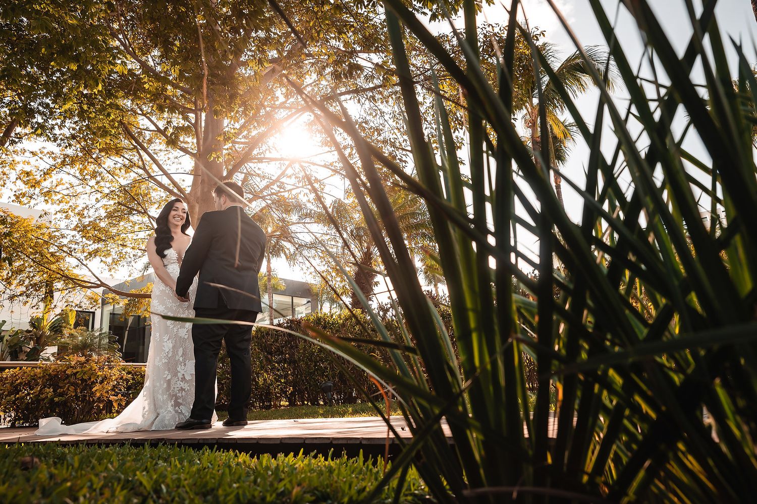 Bride and groom share a romantic moment under sunlit trees in a lush garden setting on their wedding day.