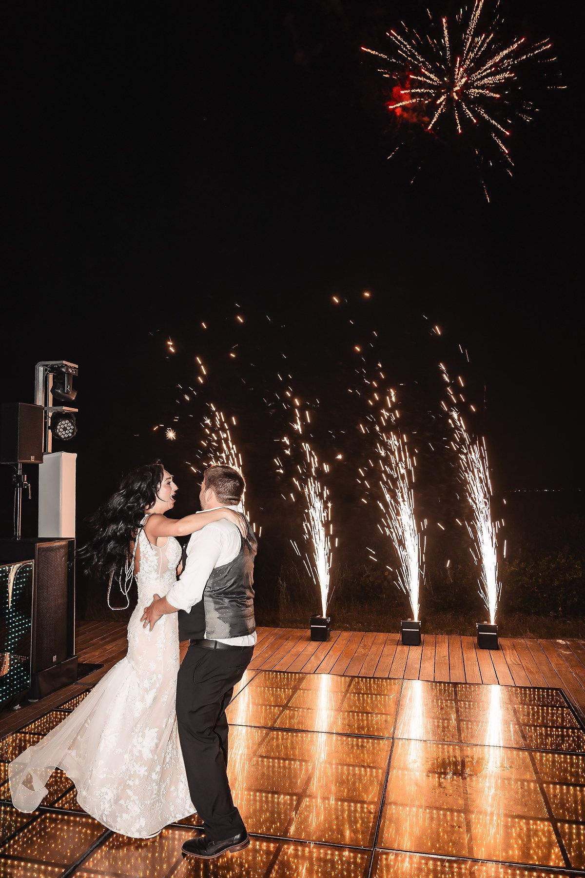 Bride and groom embrace on dance floor with sparkling fireworks display lighting up the night sky at their wedding celebration.