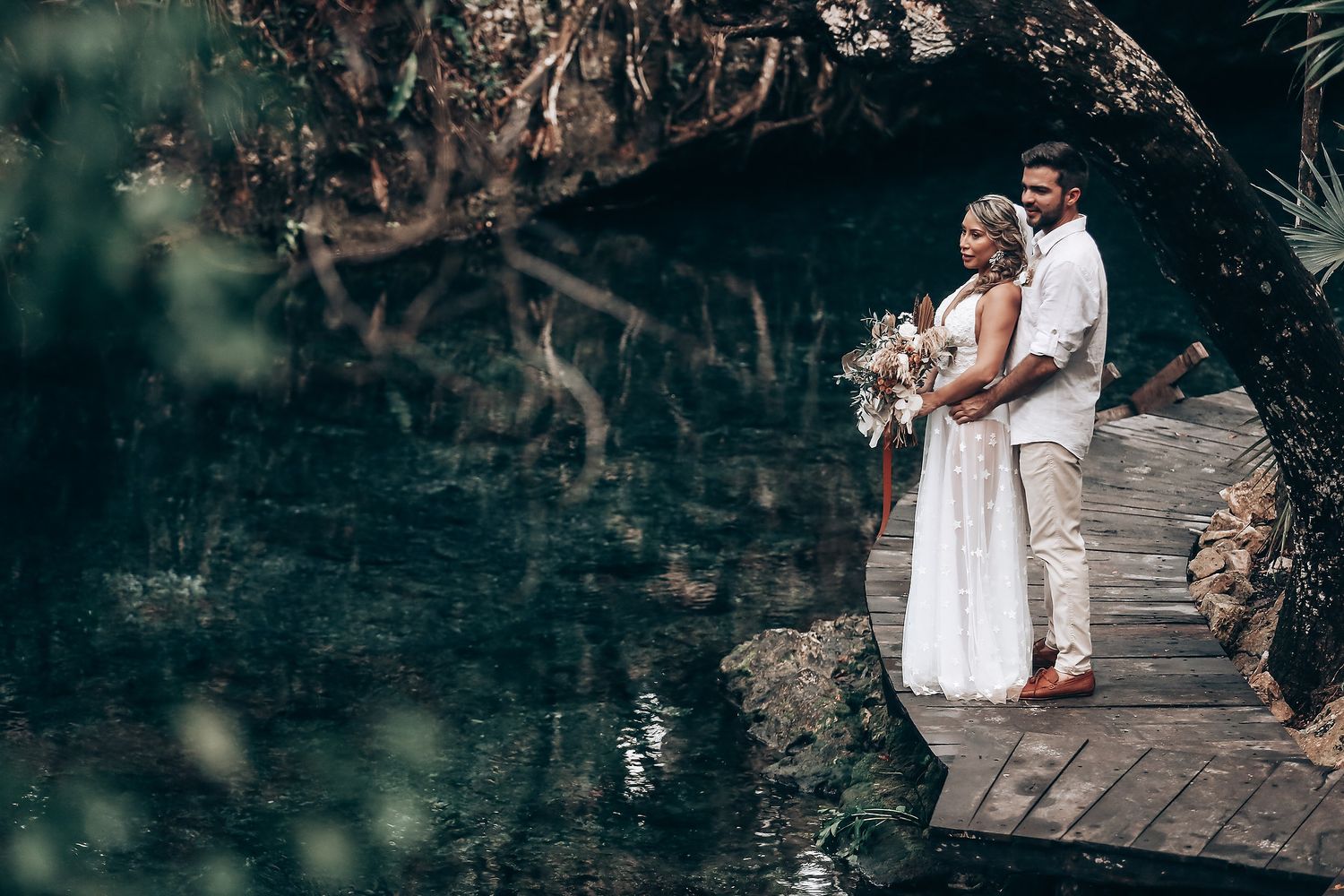 A couple stands on a wooden platform by a serene cenote surrounded by tropical foliage.