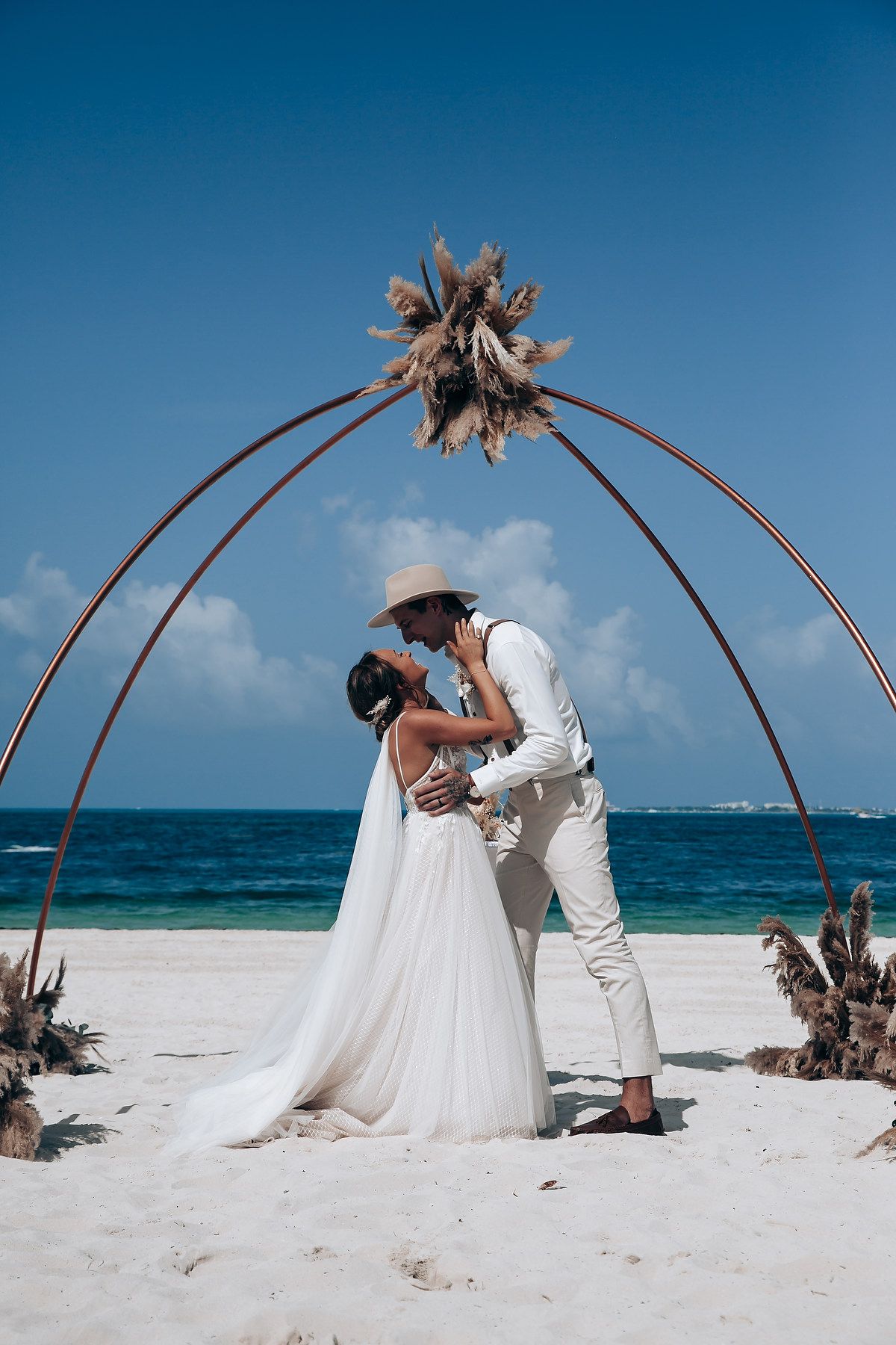 Beach wedding ceremony with curved driftwood arch and couple exchanging vows on white sand.