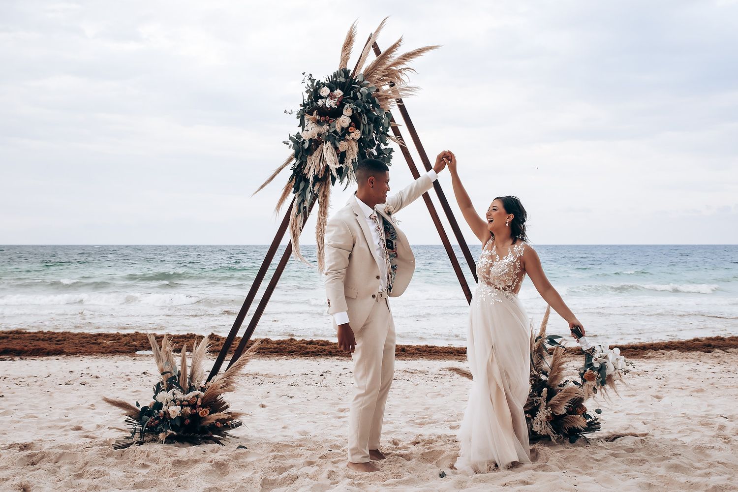 A joyful beach wedding moment under a triangle arch with ocean waves in the background.