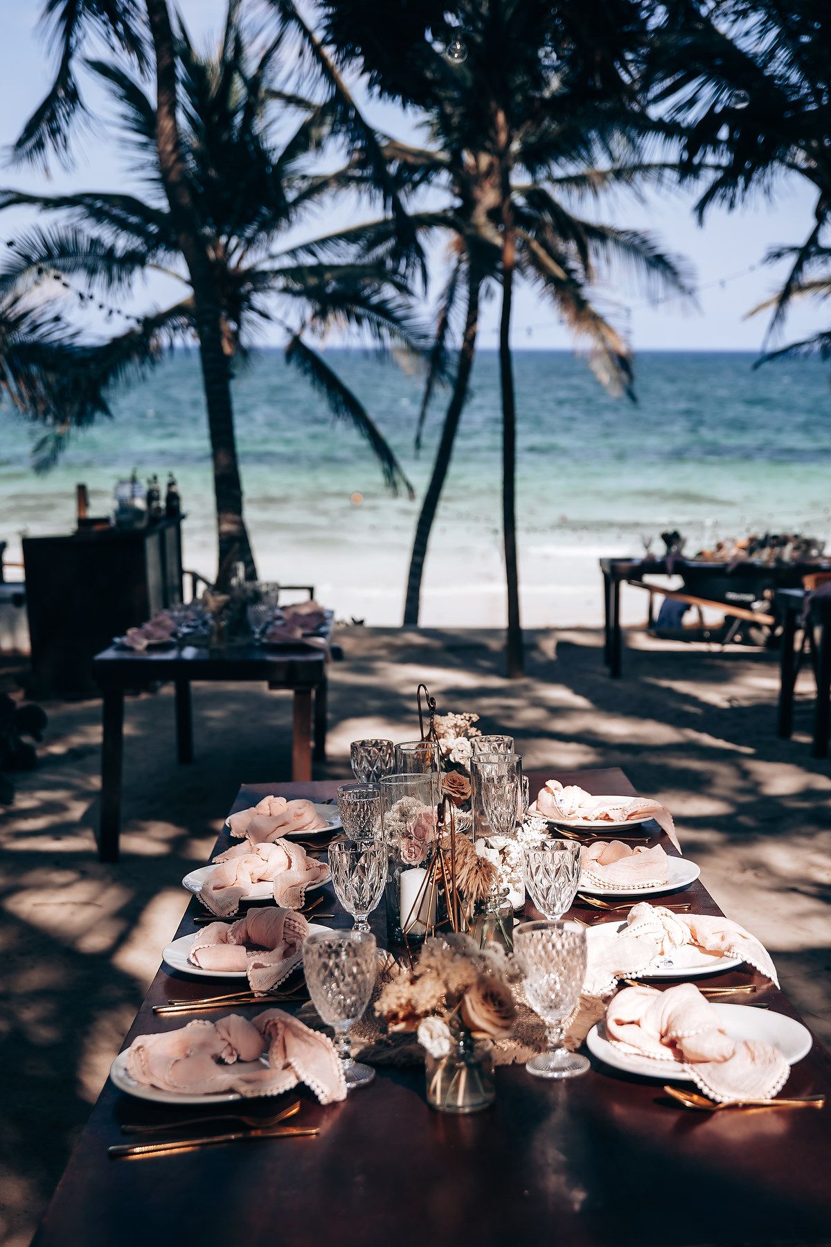 Beachside wedding reception table setup with crystal glassware under swaying palm trees.