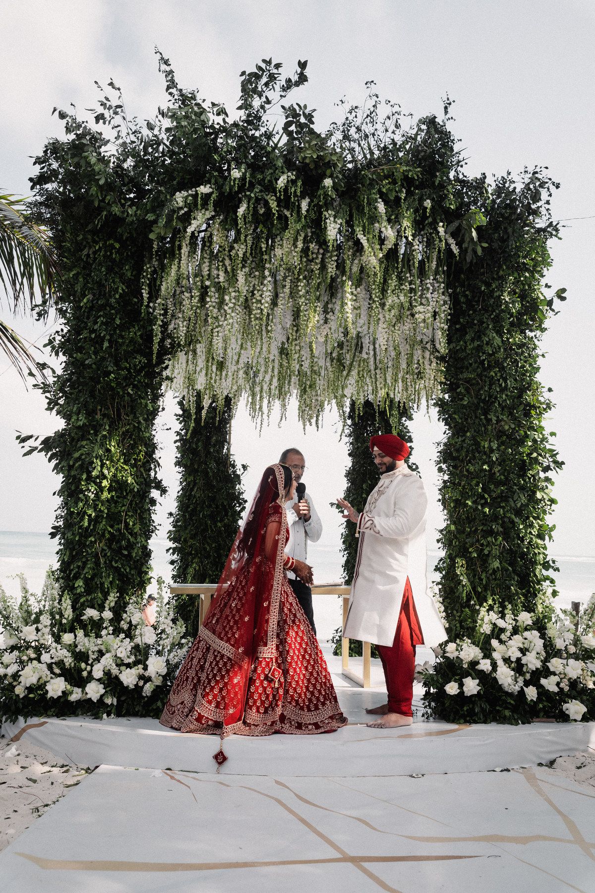 Indian bride and groom in traditional red and white attire exchange vows under green floral arch on beach.