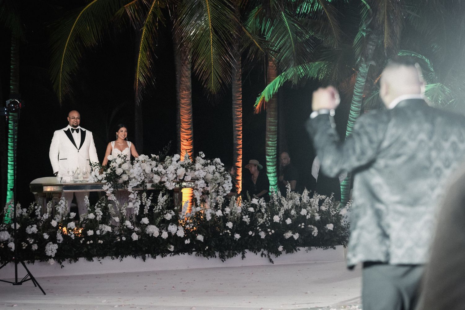 Bride and groom stand at floral-decorated altar as guest dances joyfully at tropical beach wedding.