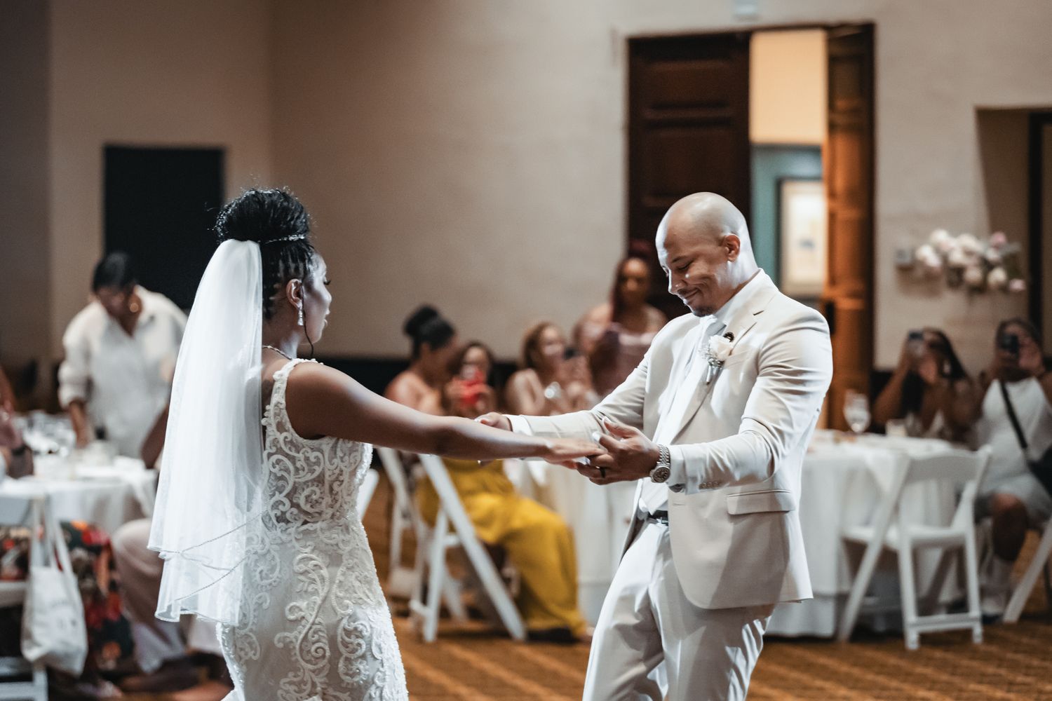 Bride and groom share their first dance at an elegant wedding reception, guests seated at white-draped tables in background.
