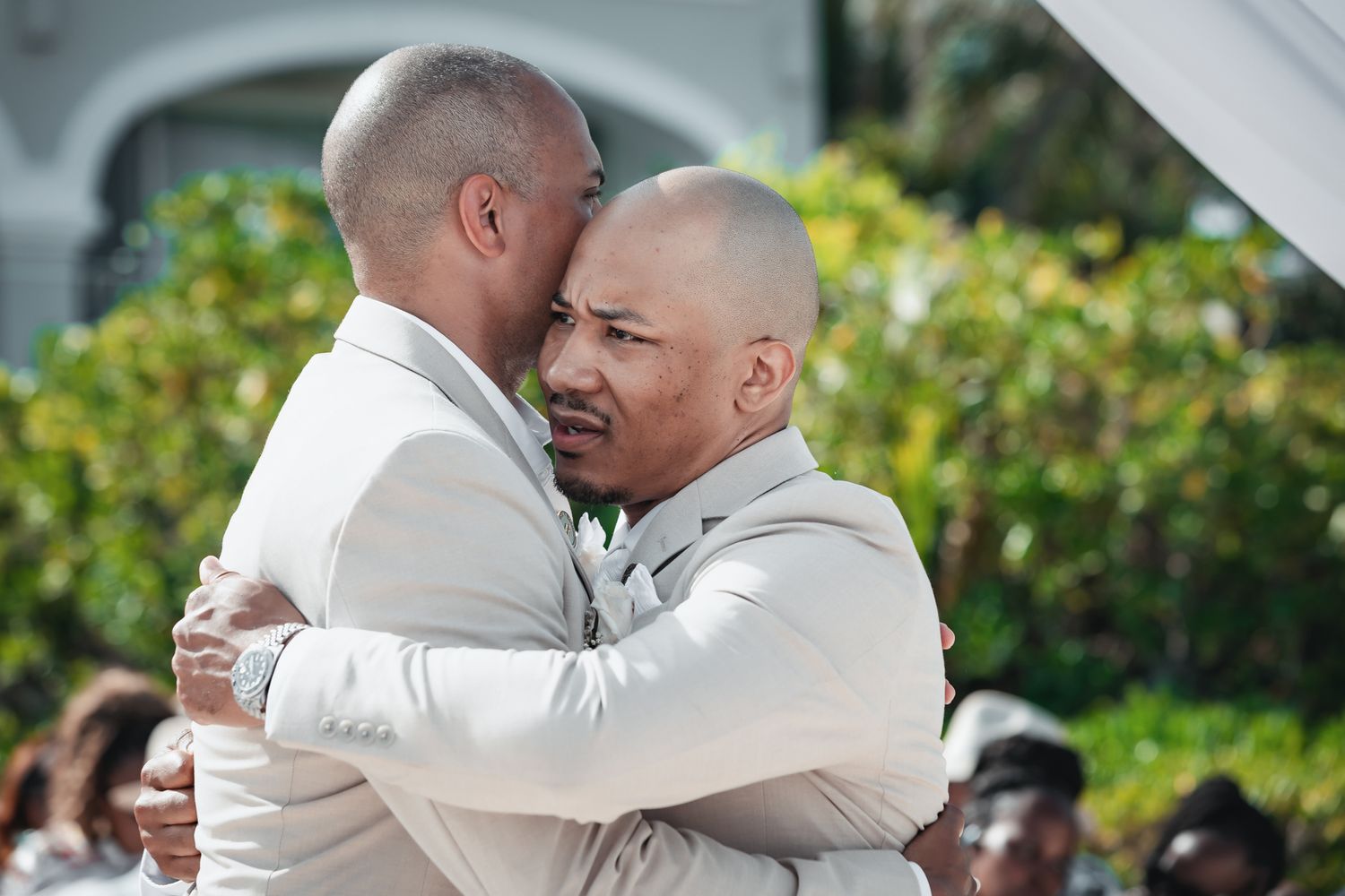 Two grooms in white suits sharing an emotional embrace at their outdoor wedding ceremony.