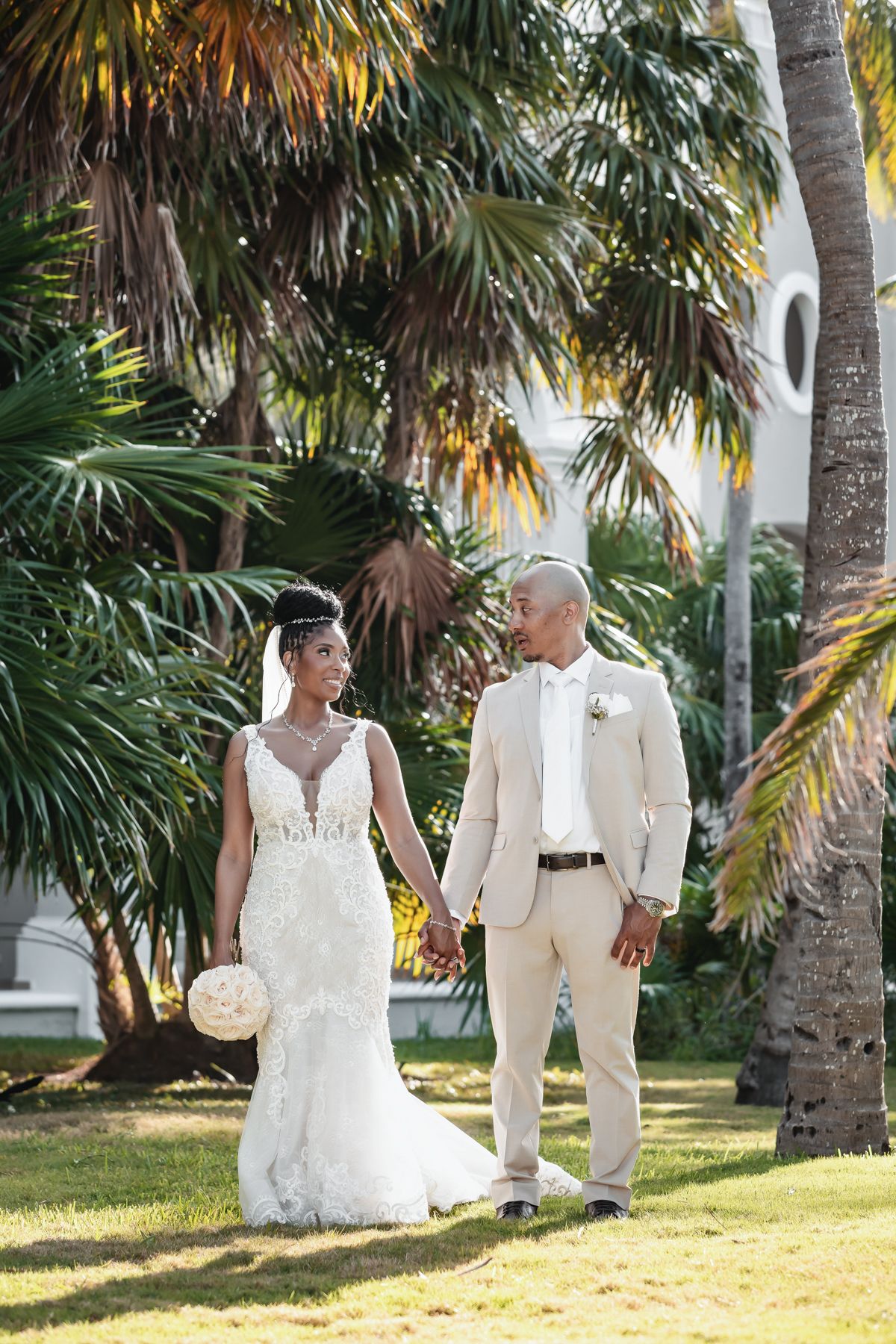 Newlyweds hold hands in tropical garden, bride in lace gown and groom in beige suit.
