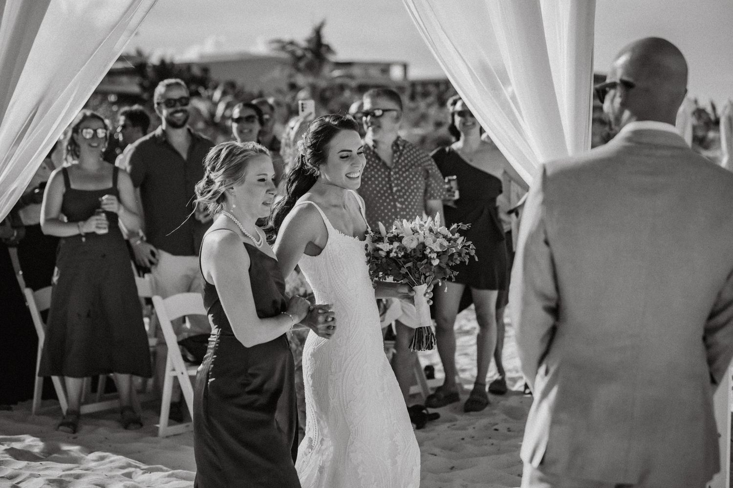 Black and white beach wedding photo of smiling bride walking down the aisle with bridesmaid holding bouquet.
