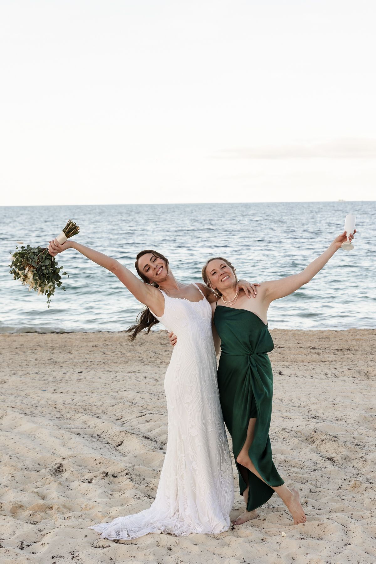 Bride in white lace gown and bridesmaid in green dress celebrate joyfully on sandy beach, arms raised holding bouquet.