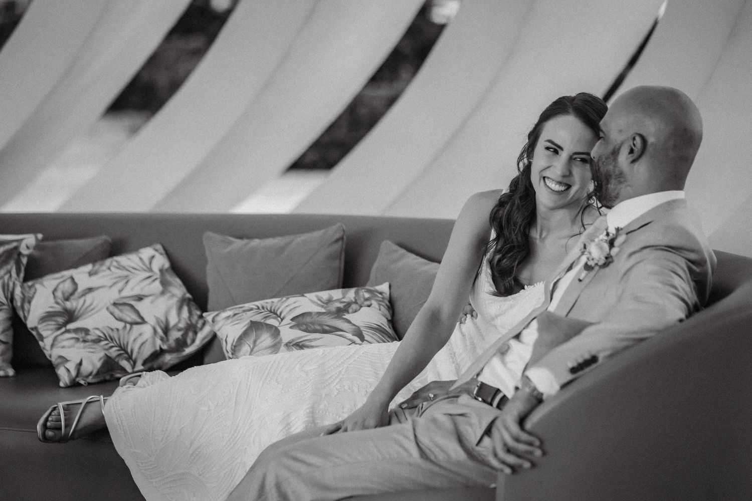 Black and white photo of a smiling bride and groom sitting together on a couch at their wedding reception.