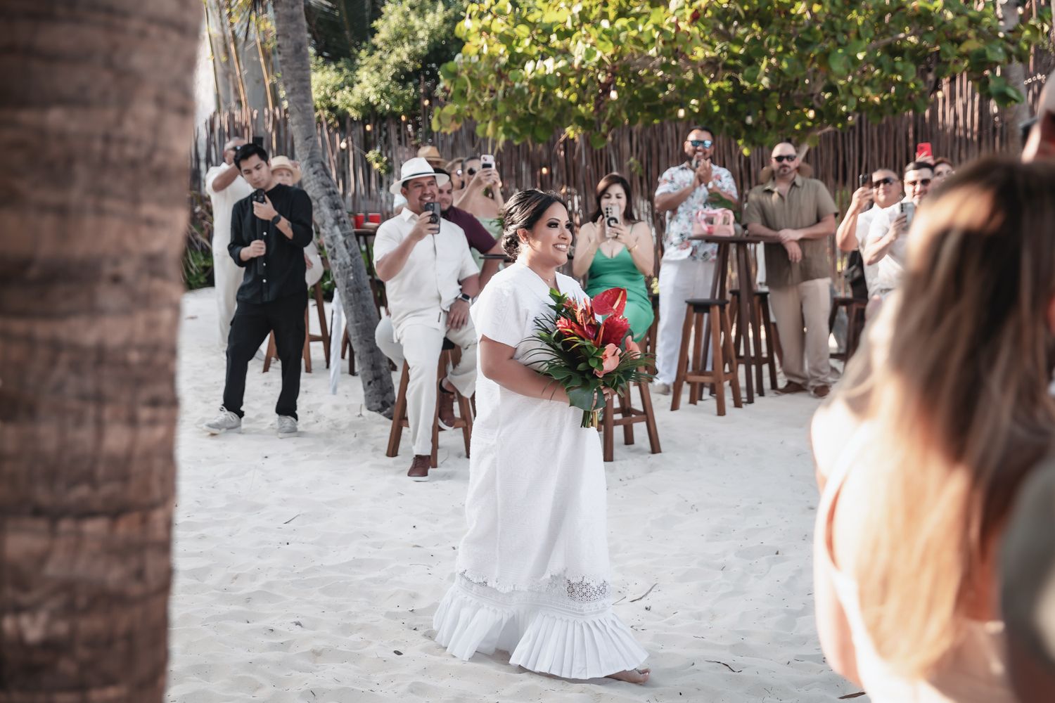 Bride in white dress walks on sandy beach holding tropical bouquet during outdoor wedding ceremony with guests watching.