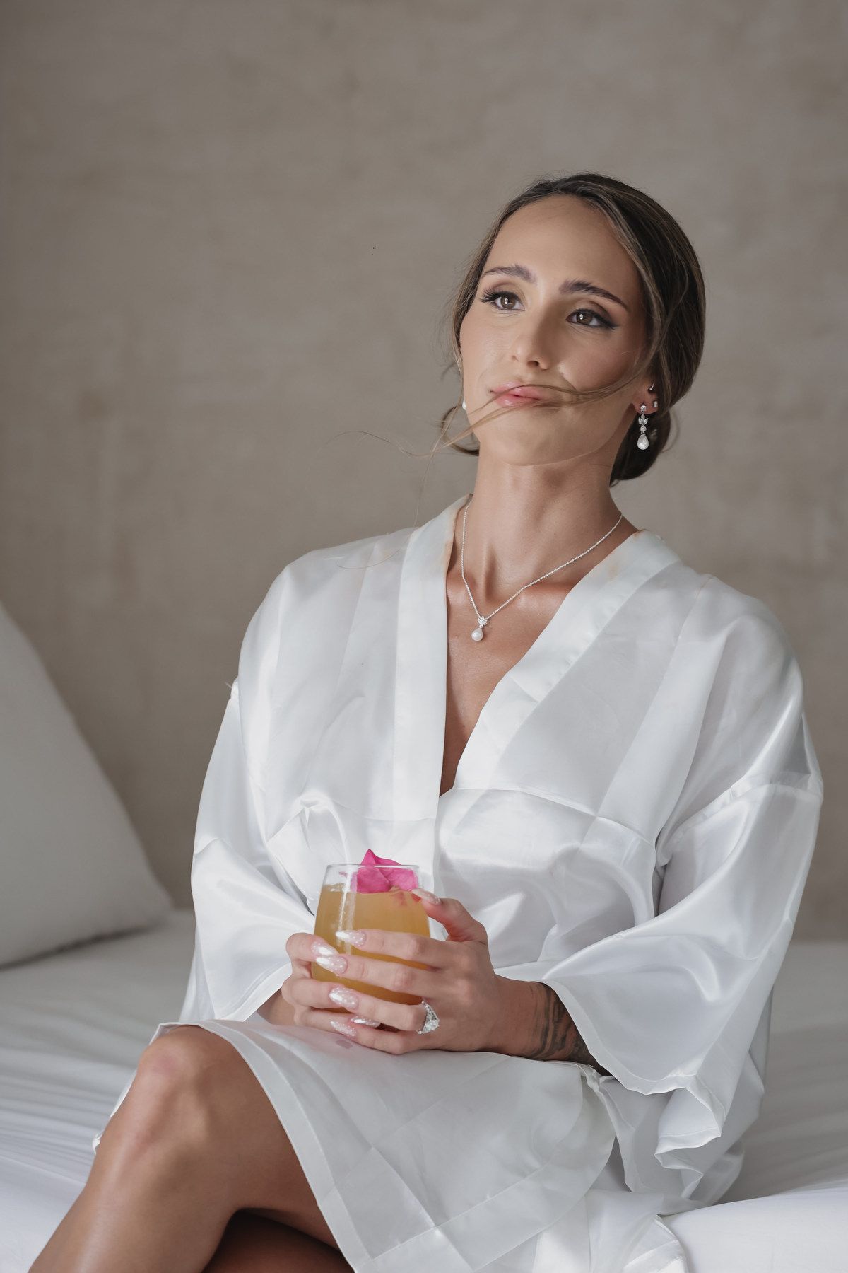 Elegant bride in white silk robe sitting on bed holding champagne glass with pink flower, wearing pearl jewelry.