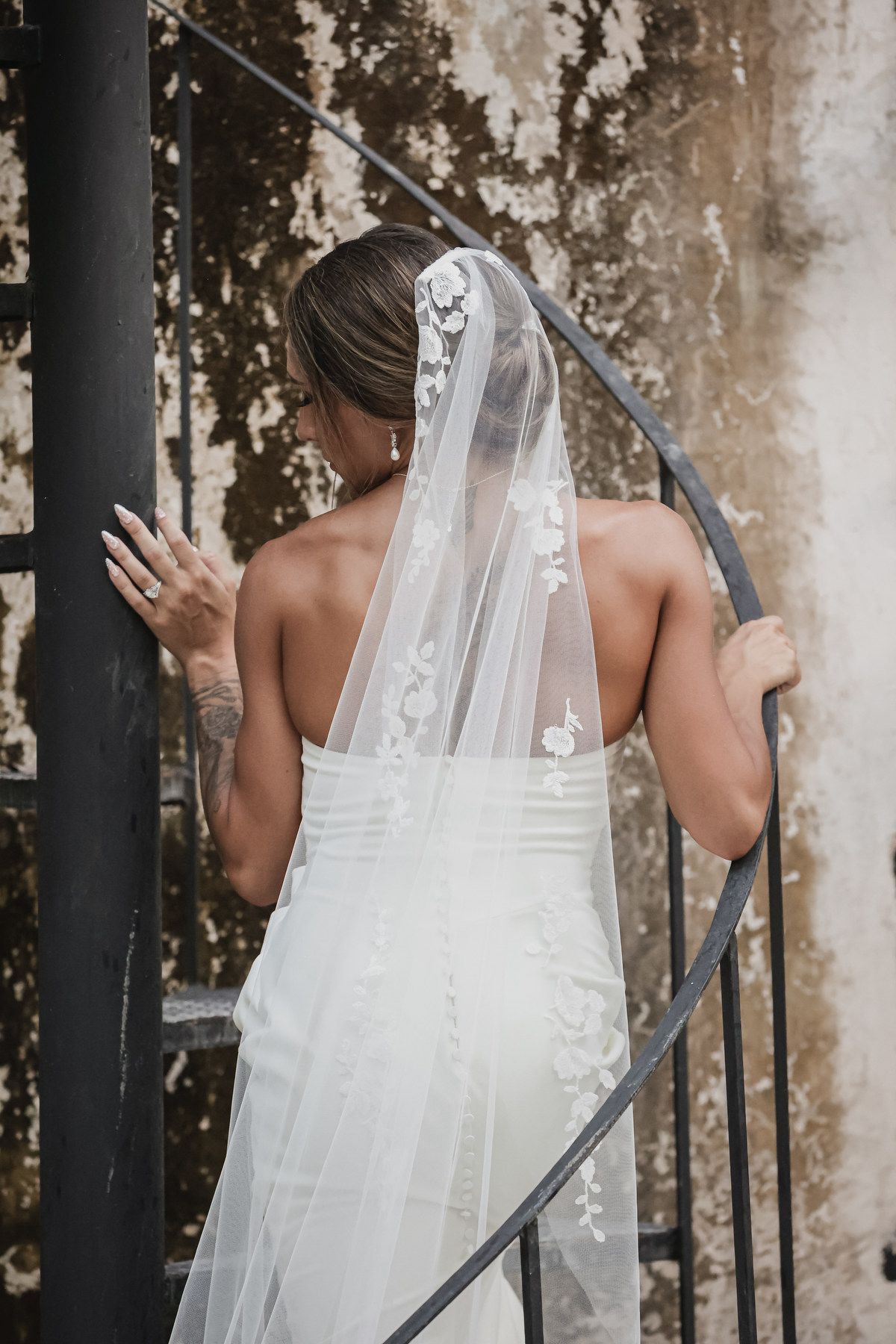 Bride in white lace wedding gown and floral veil ascending spiral staircase, viewed from behind.
