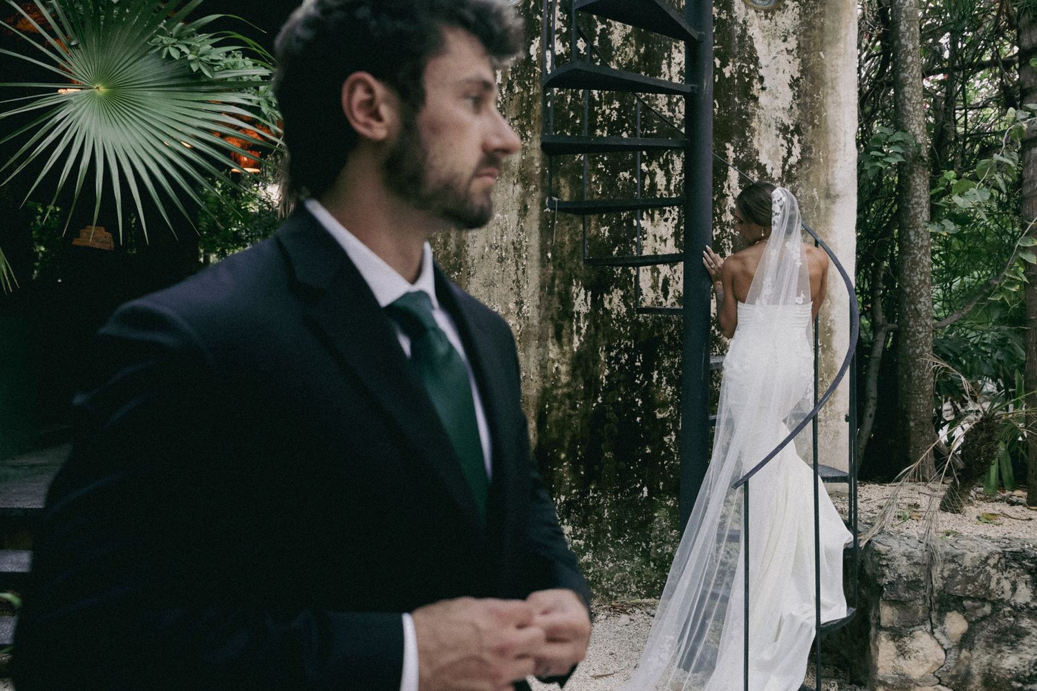 A groom in a dark suit stands outdoors as a bride in a white gown and veil poses near stone ruins.