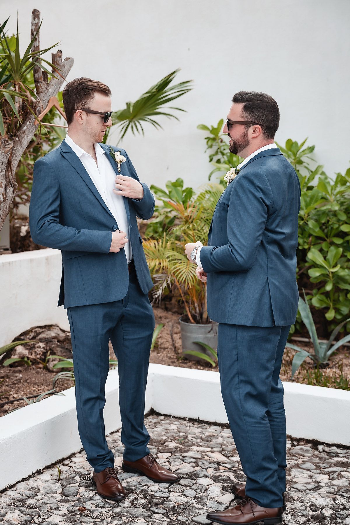 Two groomsmen in matching blue suits and sunglasses share a moment outdoors at a wedding venue with tropical greenery.