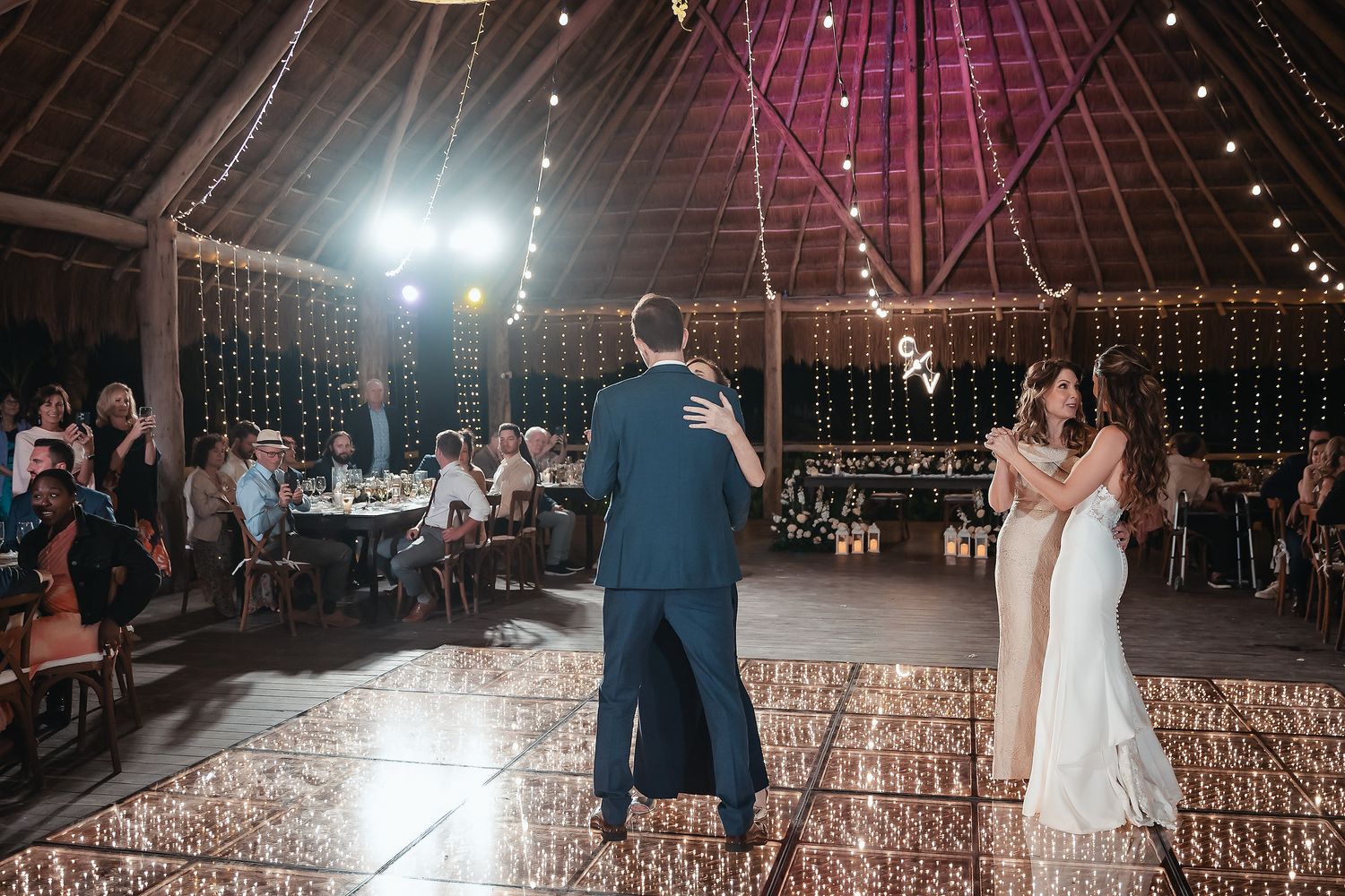 Bride and groom share first dance on glowing dance floor inside rustic barn wedding venue with string lights.