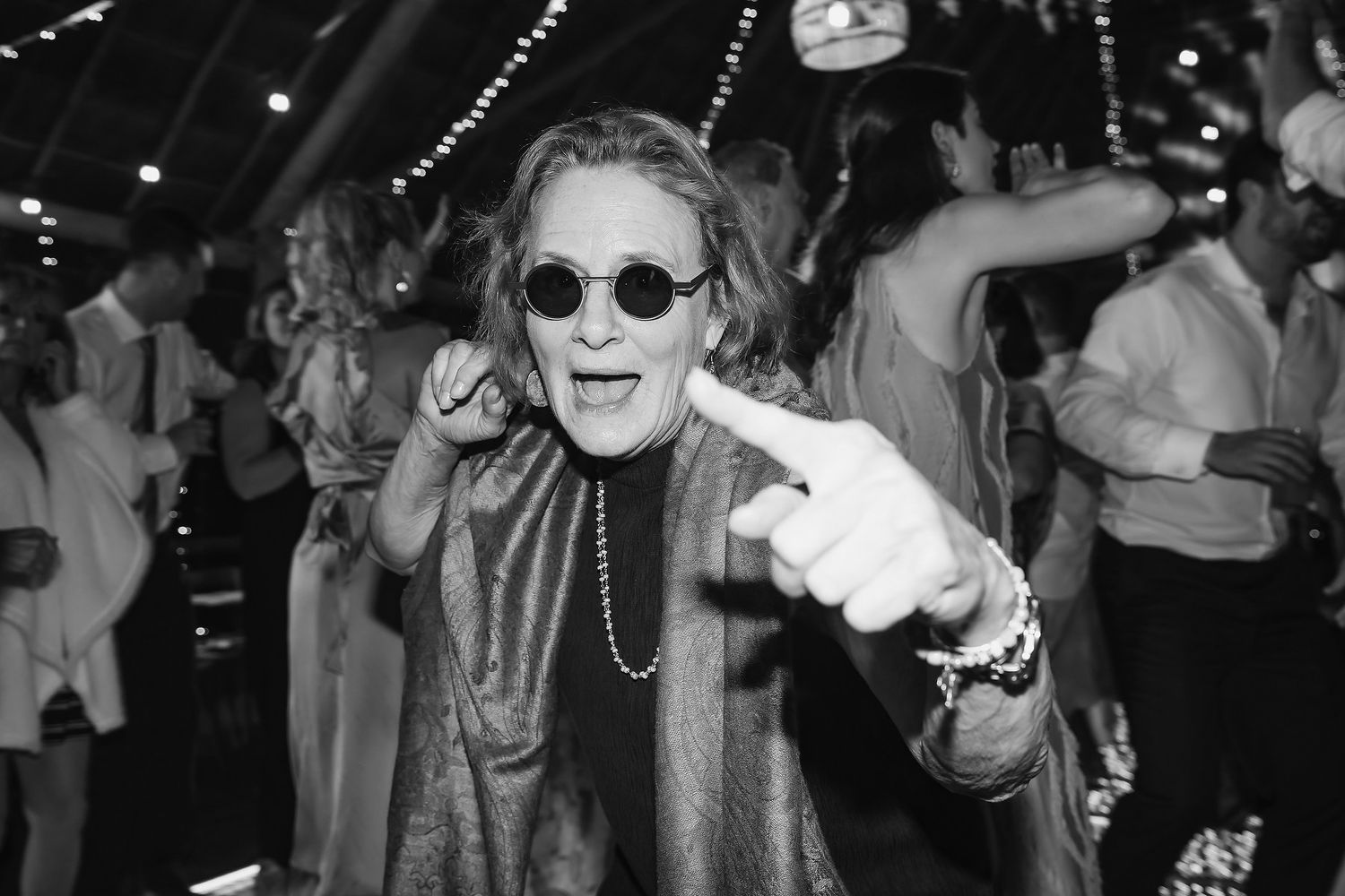 Elderly woman dancing joyfully at a party, wearing sunglasses and pearl necklace, black and white photo.