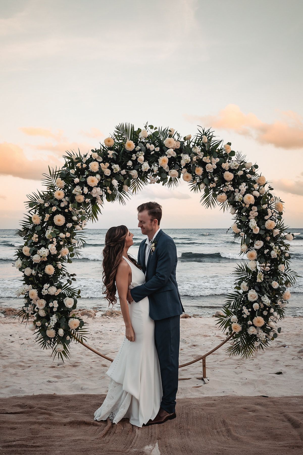 Bride and groom embrace under a floral arch on a beach at sunset, surrounded by white roses and tropical greenery.