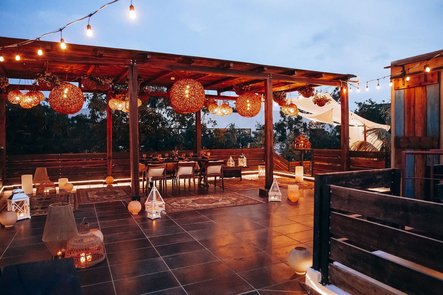 Rustic outdoor wedding venue terrace with glowing wicker lanterns, string lights, and candles at dusk.