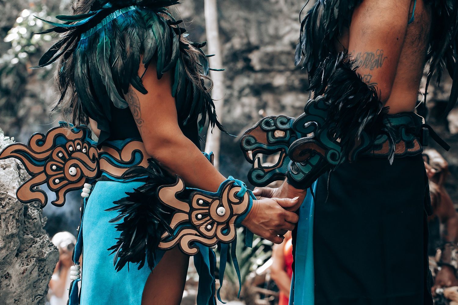 Close-up of two Mayan ceremonial dancers in turquoise and black feathered costumes with intricate serpent decorations.